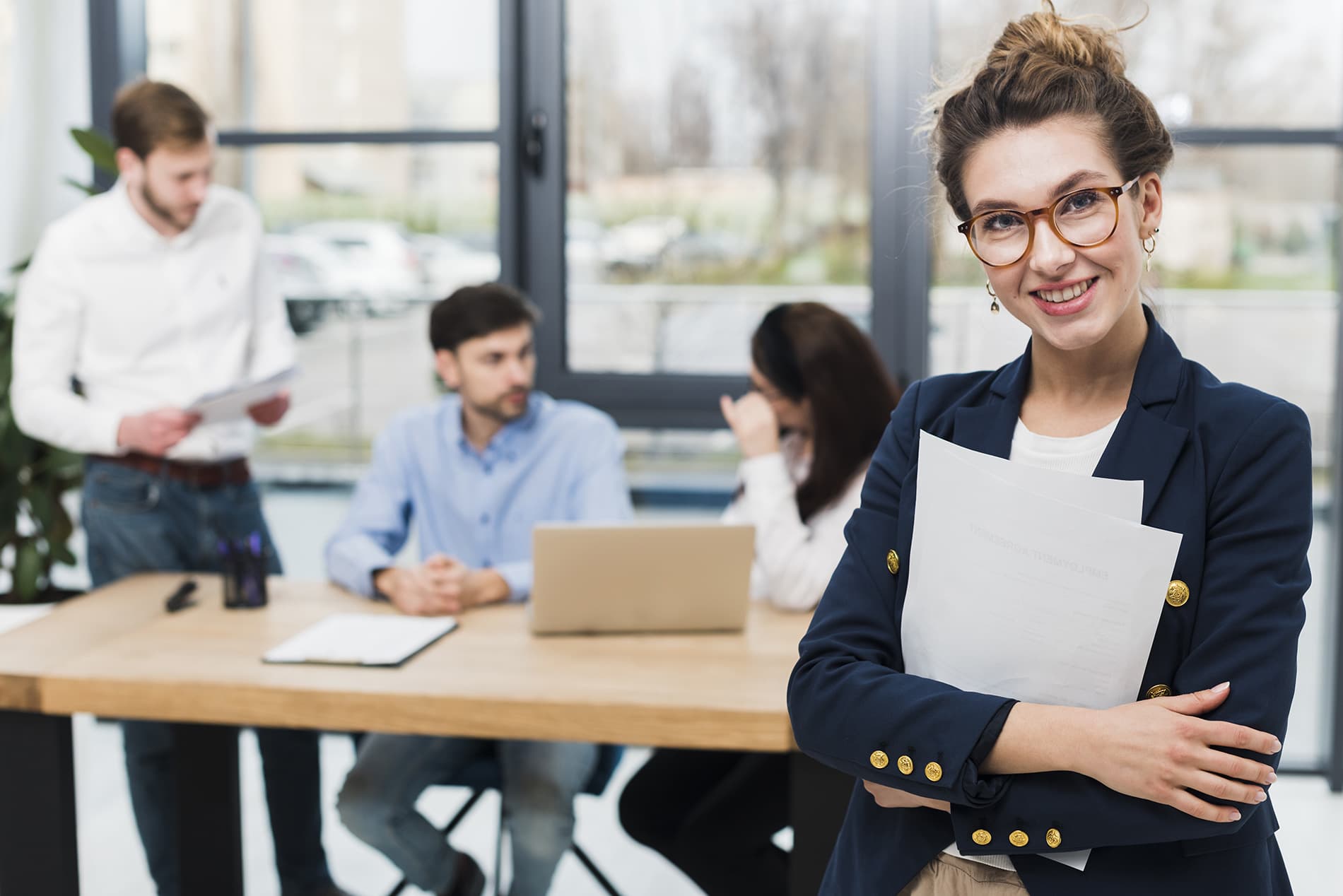 Woman in a board room holding documents while coworkers in the background are having a meeting.